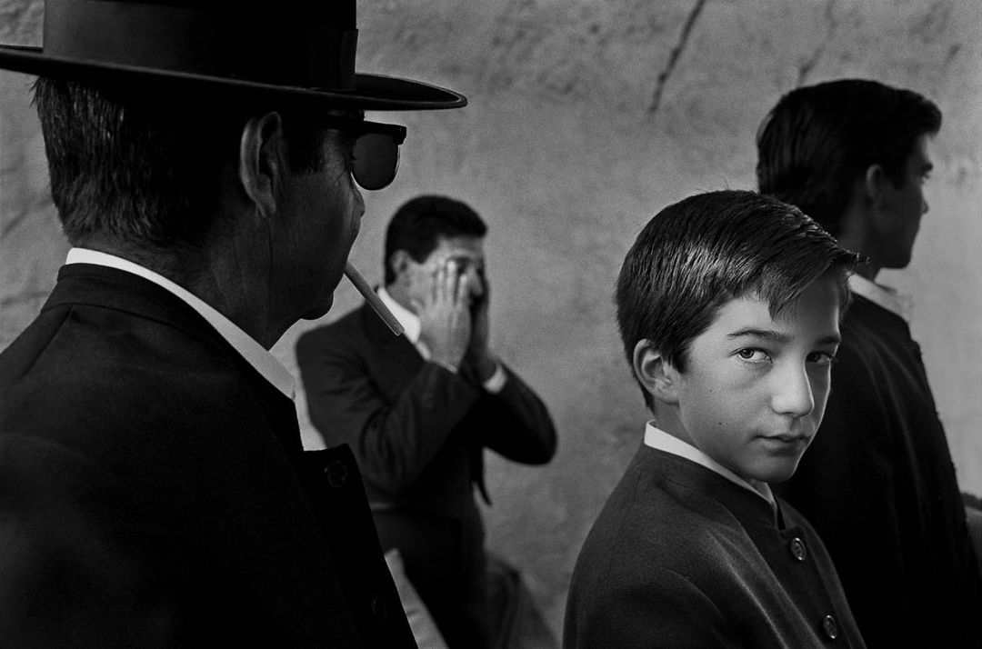 Black and white photo of a young boy surrounded by adults in church. The boy is giving a mischievous glance. Taken from Johan Willner's collection Wind Upon the Face of Waters, photographed during the seventies and eighties.