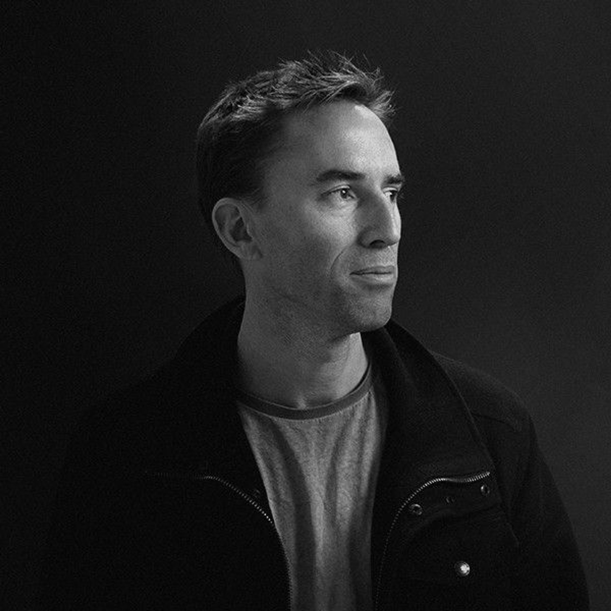 A black-and-white photo of Matt, wearing a t-shirt and jacket. Soft light is shining on him from one side, and his face is turned toward it. He’s got a small smile on his face.