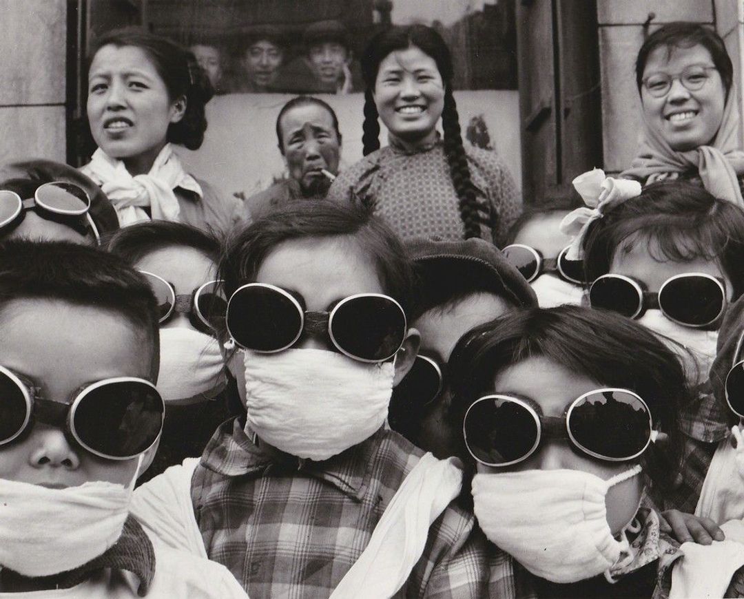 Vintage silver print taken by Agnes Varda of a bunch of Chinese children wearing goggles and masks to prepare to spend time in the Gobi desert. Adults standing behind them, perhaps teachers or parents.