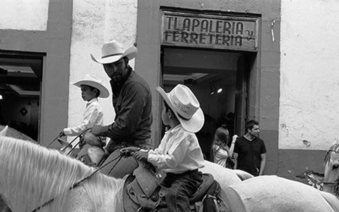An adult male cowboy on a horse, talking to a child cowboy on another horse. A sign behind them says "Apaleria Ferreira".