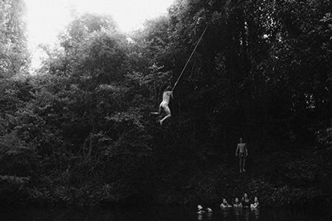 A nude person swinging off a tree by a rope, ready to let go and fall into the water below.