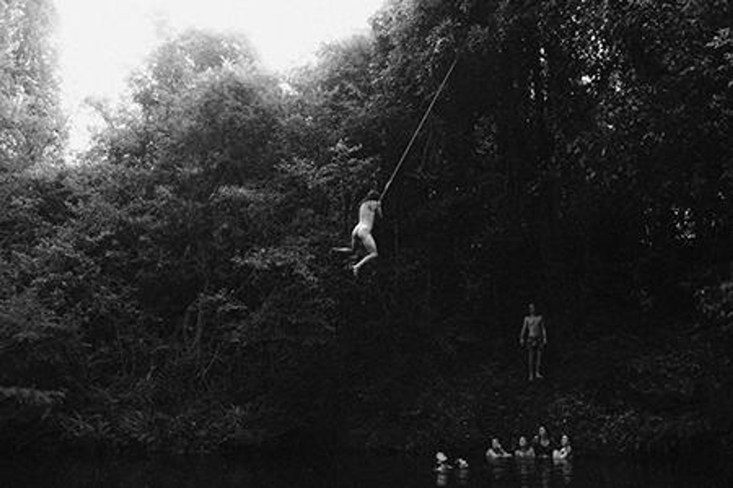 A nude person swinging off a tree by a rope, ready to let go and fall into the water below.