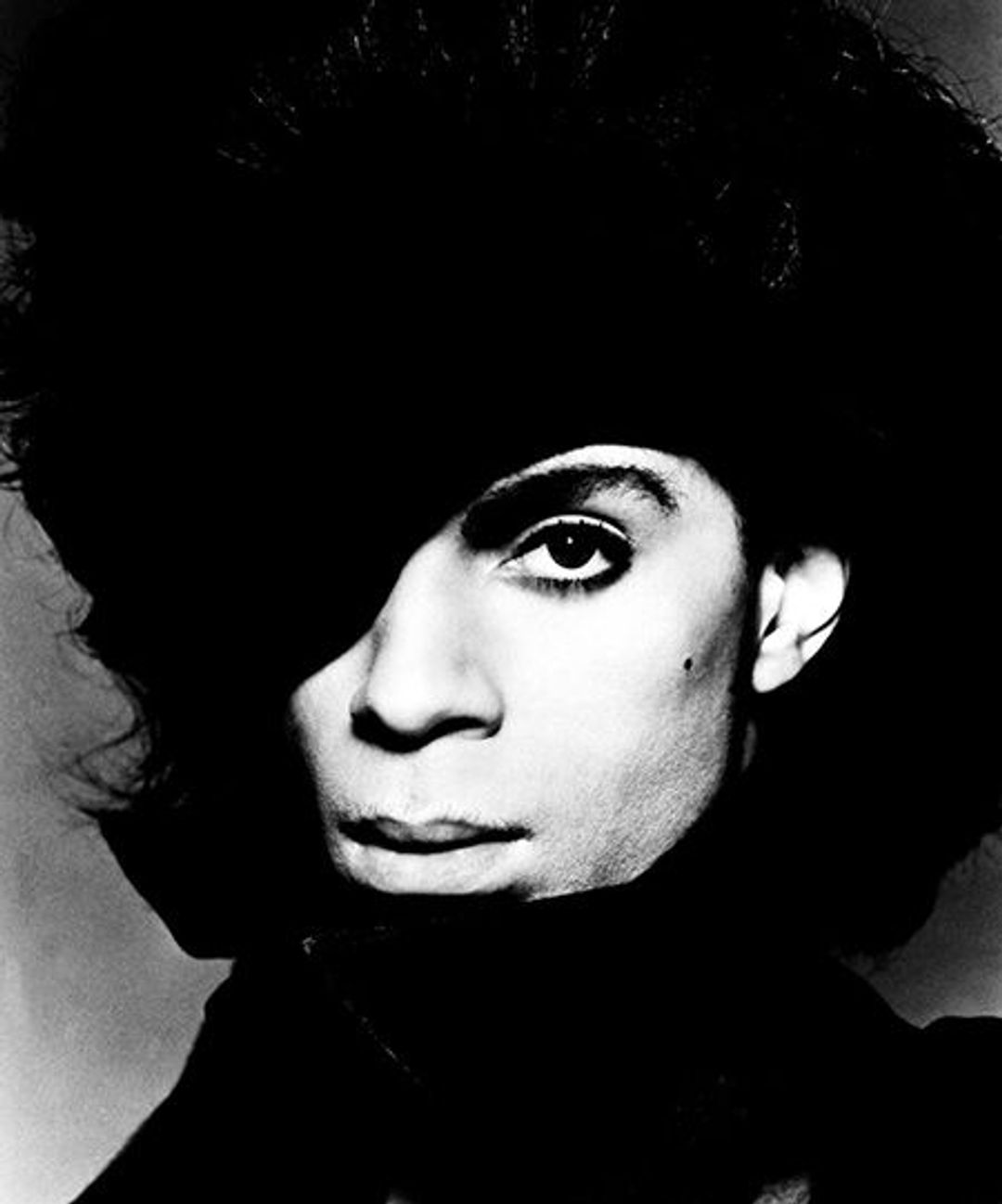 A heavily contrasted photo of Prince's face. A mole on his cheek. Stone cheekbones. He's possibly wearing a large black hat but the light's too dark to see.