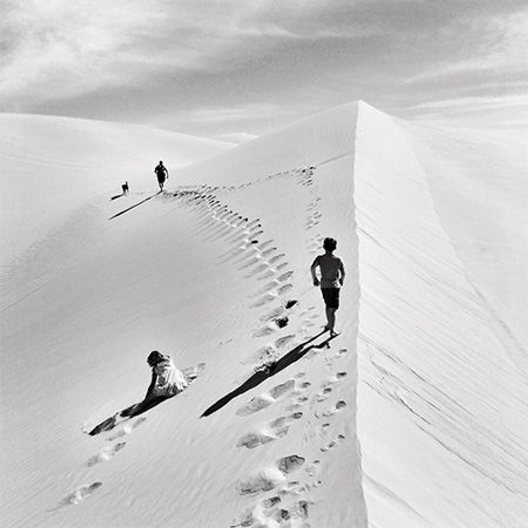 Kids running through the desert in the daytime atop a sand dune. The sand dune acts as the camera line into the distance. There's a little girl off to the left who is sitting a little way down the dune, as if exhausted.