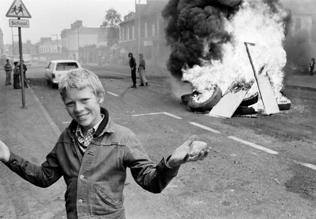 Photo of white kid standing and smiling in front of a fire containing wood planks and tyres. Shrugging with his hands up. By Chris Steele Perkins.