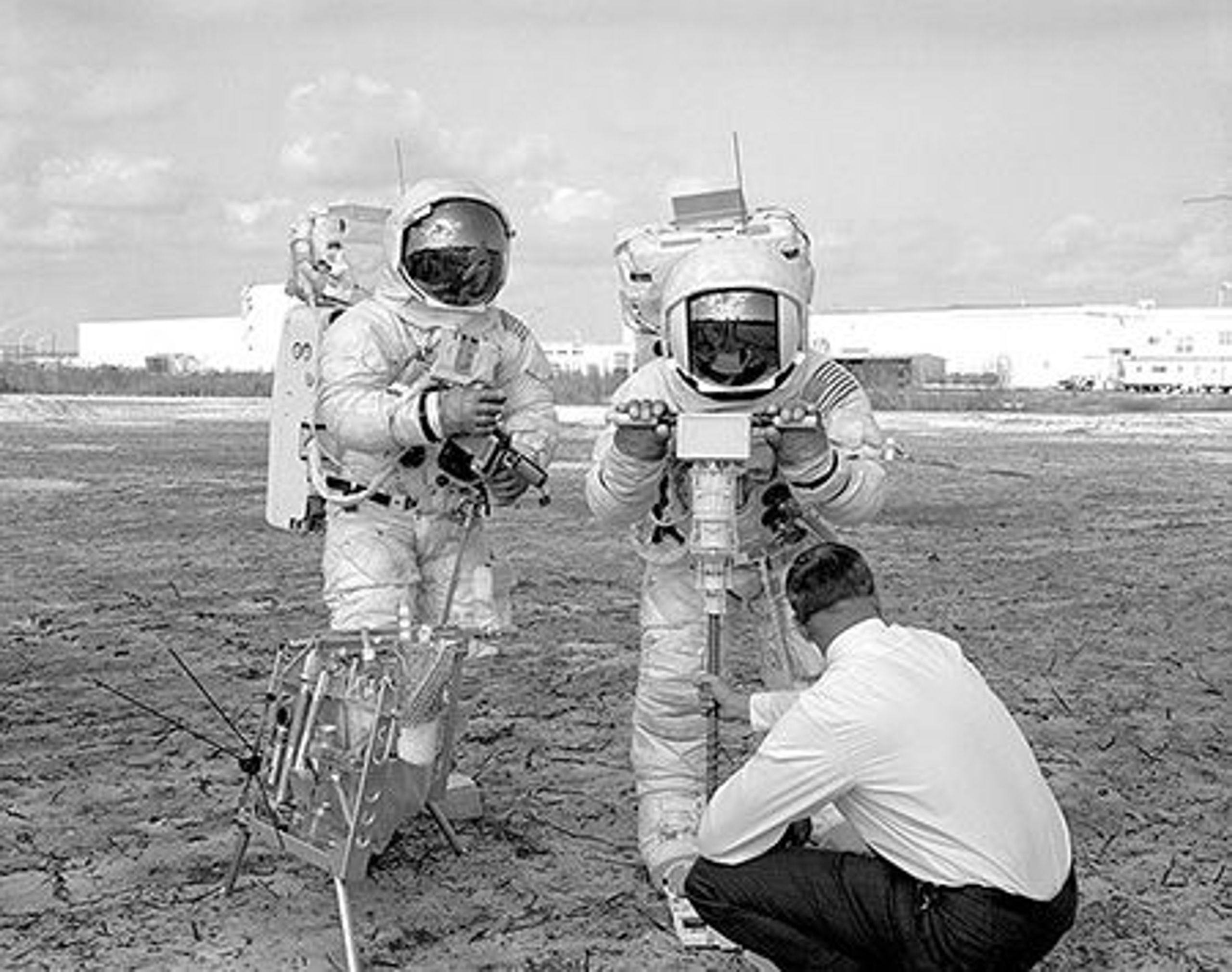 Astronauts in front of a warehouse, and a man in a suit adjusting their equipment. They have a big white camera pointing at the ground.